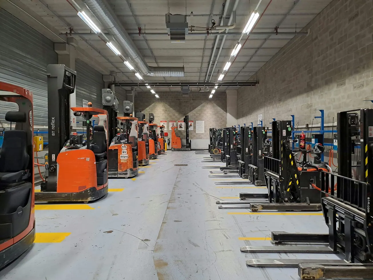 A warehouse interior with rows of parked orange and black forklifts lining both sides. The space is well-lit and organized, conveying efficiency.