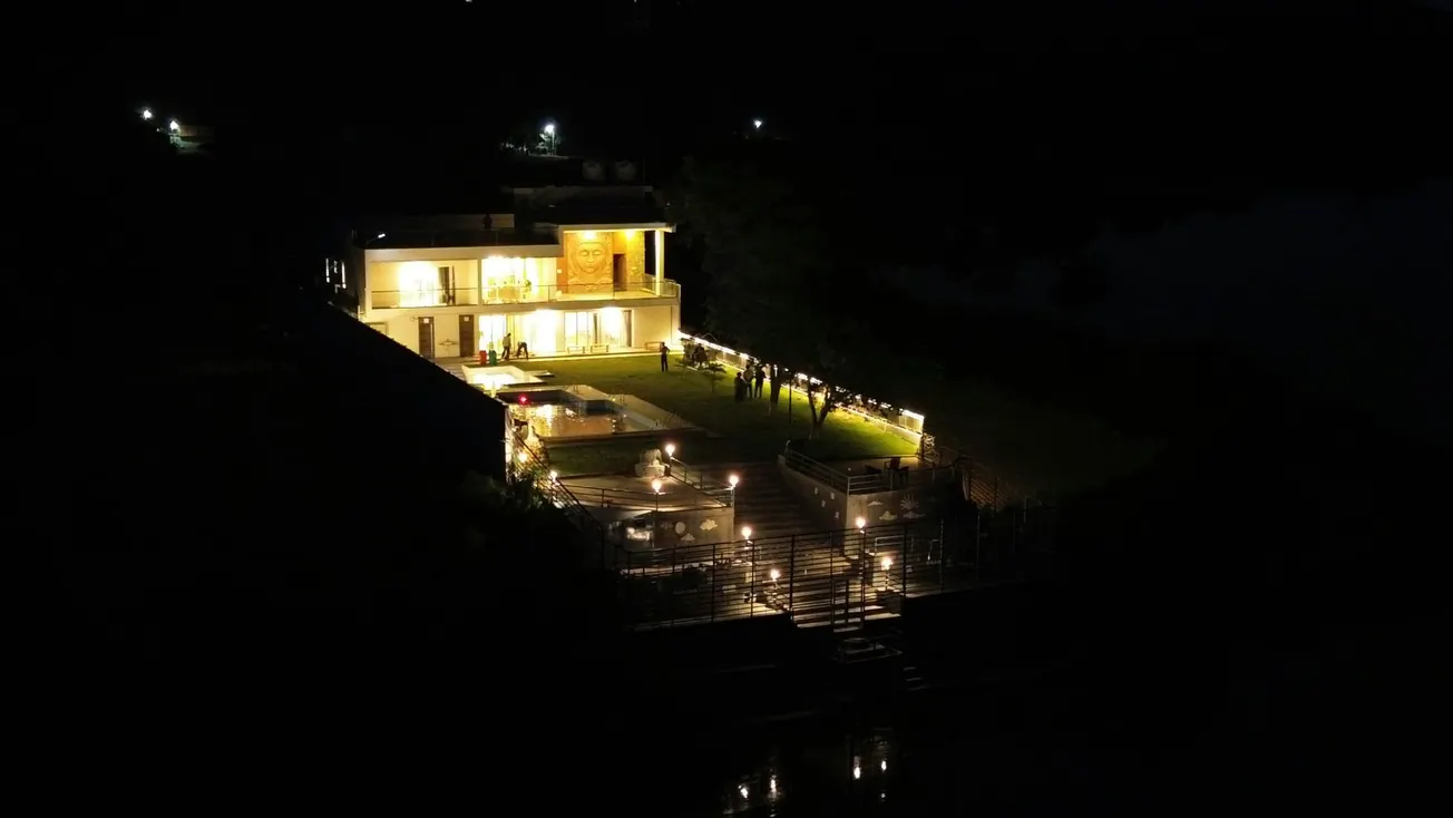 Aerial night view of a brightly lit modern house with a spacious lawn. Warm lighting creates a cozy ambiance. Groups of people gather on the lawn.