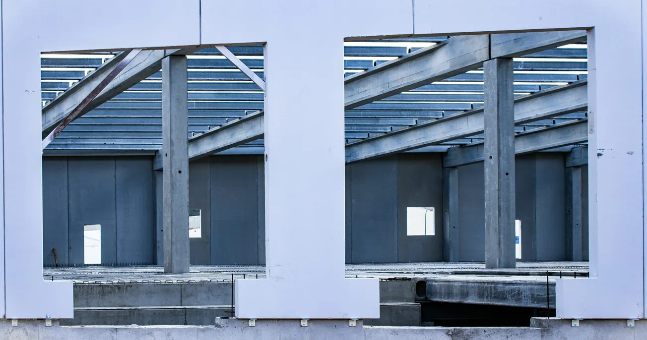 A partially constructed building with exposed steel beams and concrete pillars. Large rectangular openings reveal the structure's skeletal framework. The tone is industrial.