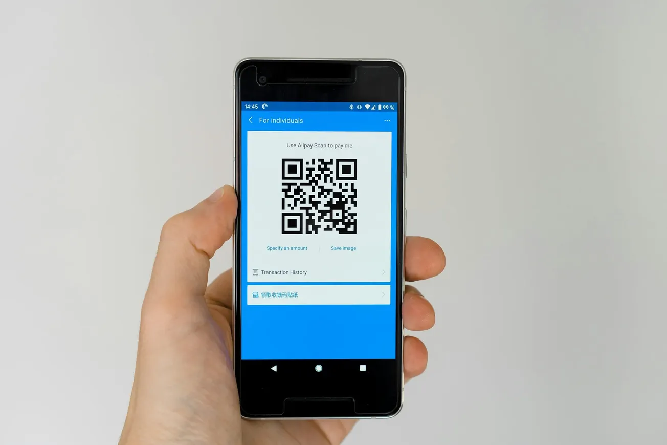 A hand holds a smartphone displaying a QR code on a blue app screen, intended for payment. The tone is efficient and modern.