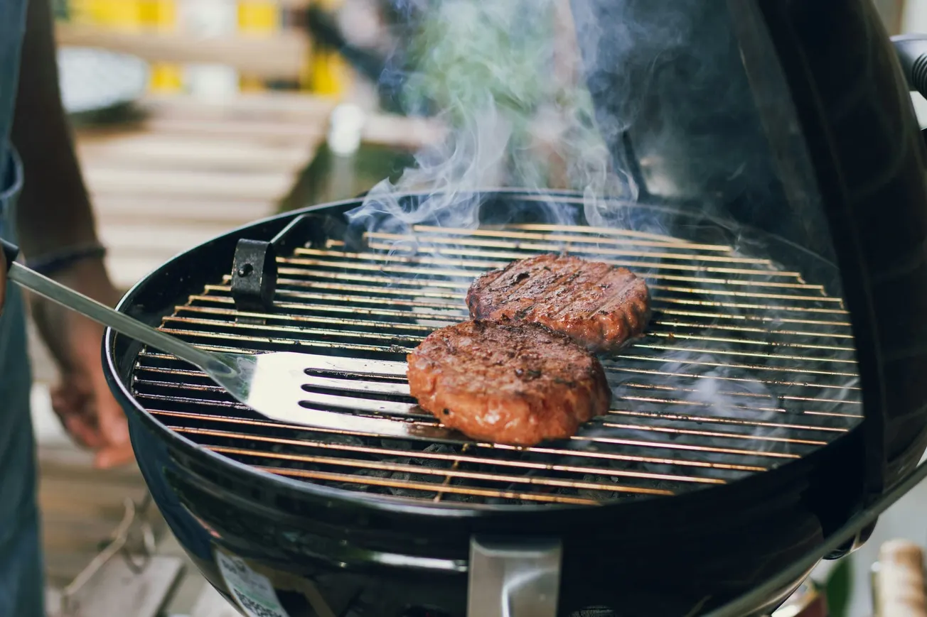 Two burger patties sizzling on a round grill with smoke rising. A spatula and a warm, inviting outdoor setting are visible in the background.