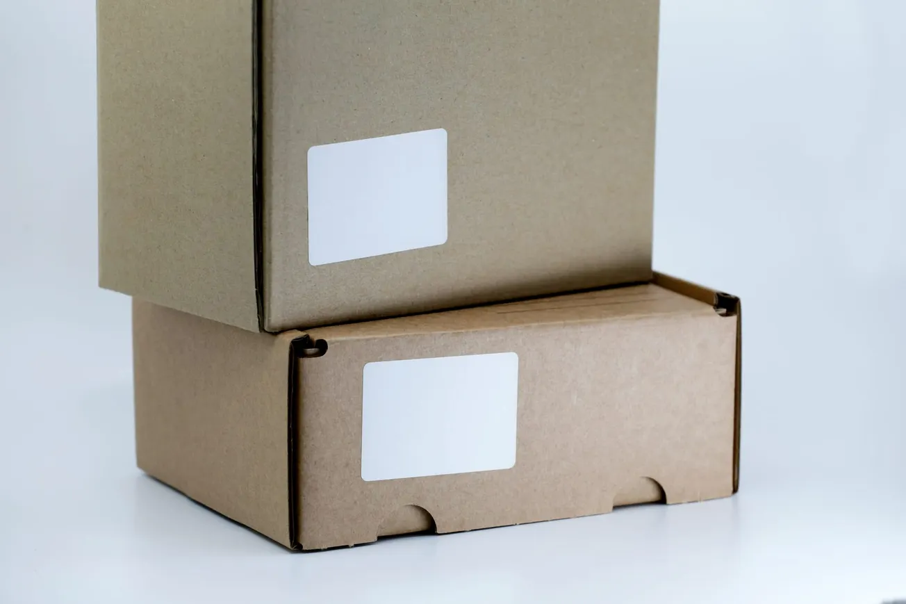 Two stacked cardboard boxes with blank white labels on a white background. The boxes are plain and have a simple, utilitarian appearance.