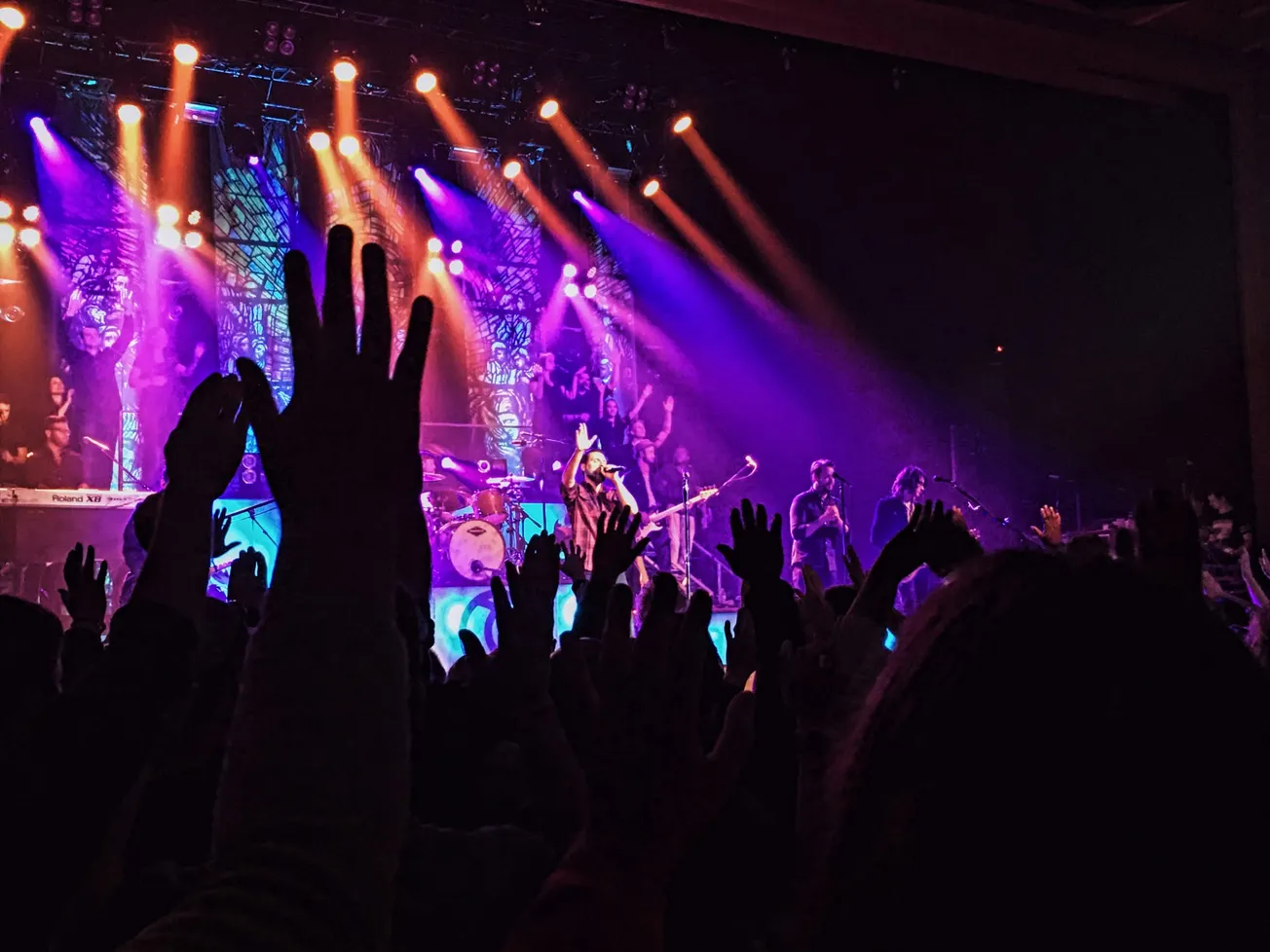 Audience with raised hands at a lively concert. Vibrant purple and orange stage lights illuminate the band performing against a vivid background. Energetic atmosphere.