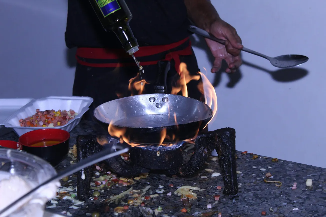 Chef pouring oil into a flaming pan on a stovetop. Nearby, bowls with chopped vegetables and sauce are ready for cooking. Dynamic and intense.