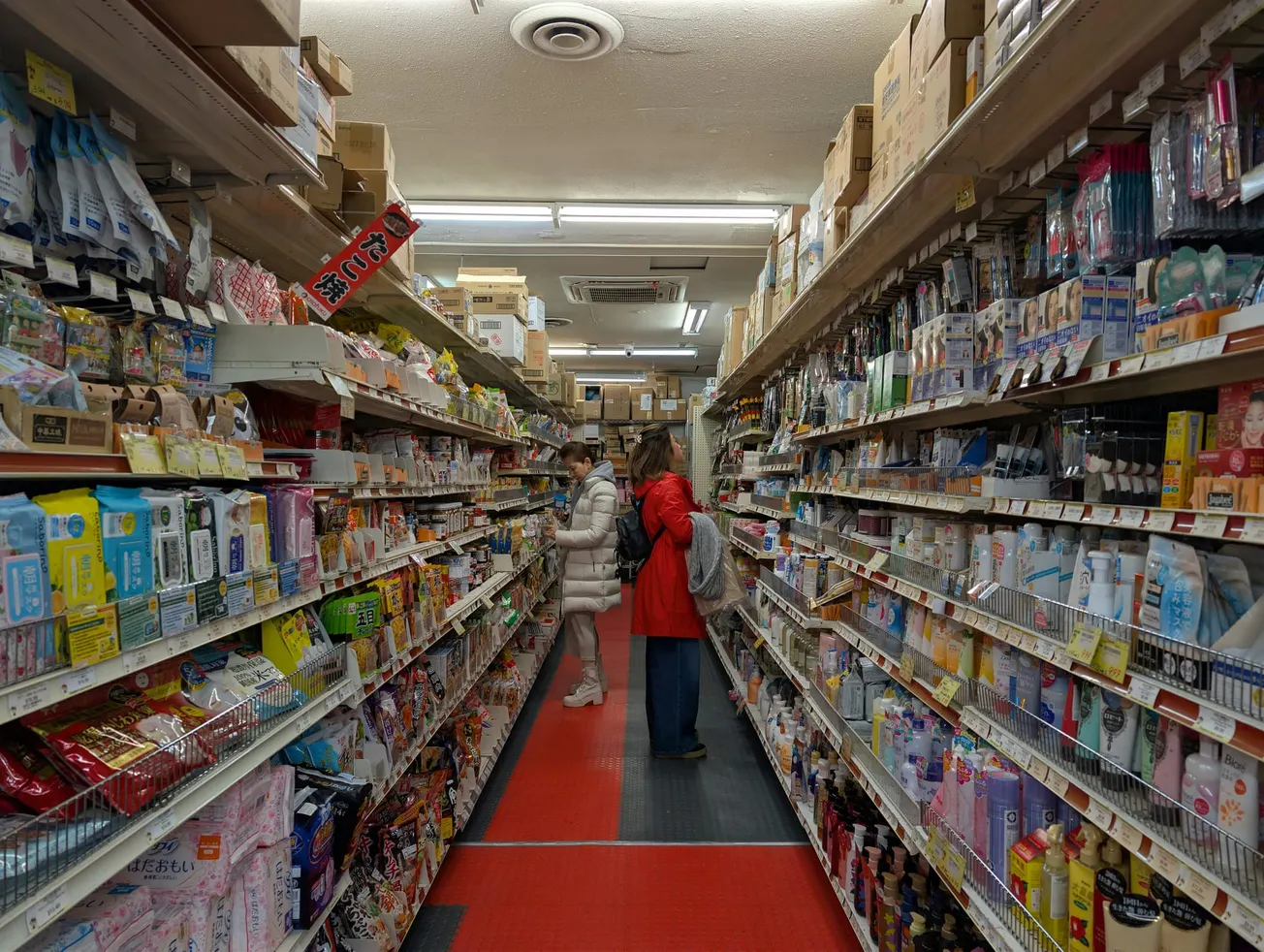 Aisle in an Asian grocery store with shelves packed with snacks and toiletries. Two people in coats browse items, creating a busy yet focused vibe.