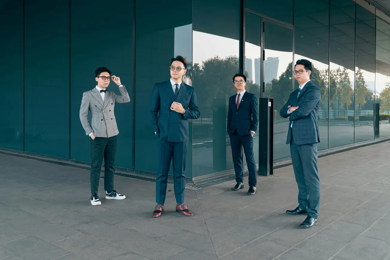 Four men in suits stand confidently outside a modern glass building. They exude a sense of professionalism and teamwork, with a backdrop of trees and city skyline reflecting in the windows.