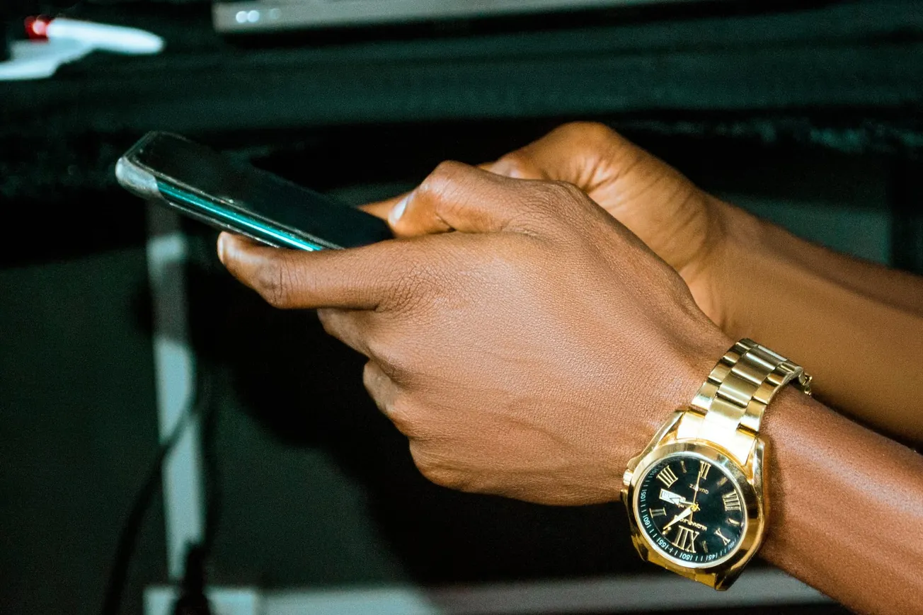 A hand wearing a gold watch with a black face holds a smartphone. The background is dim, highlighting the focus on technology and time management.