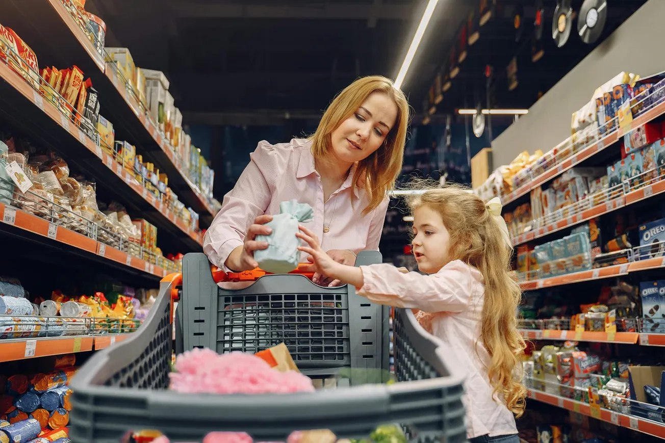 A woman and a young girl shop together in a grocery store, placing items in a cart. Shelves filled with various products line the aisle, creating a warm, familial scene.