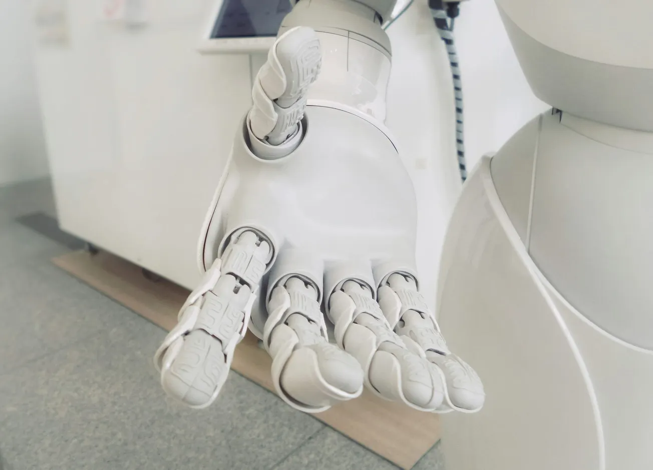A close-up of a white robotic hand reaching out, showcasing intricate joints and design. The background features a modern, minimalistic setting.