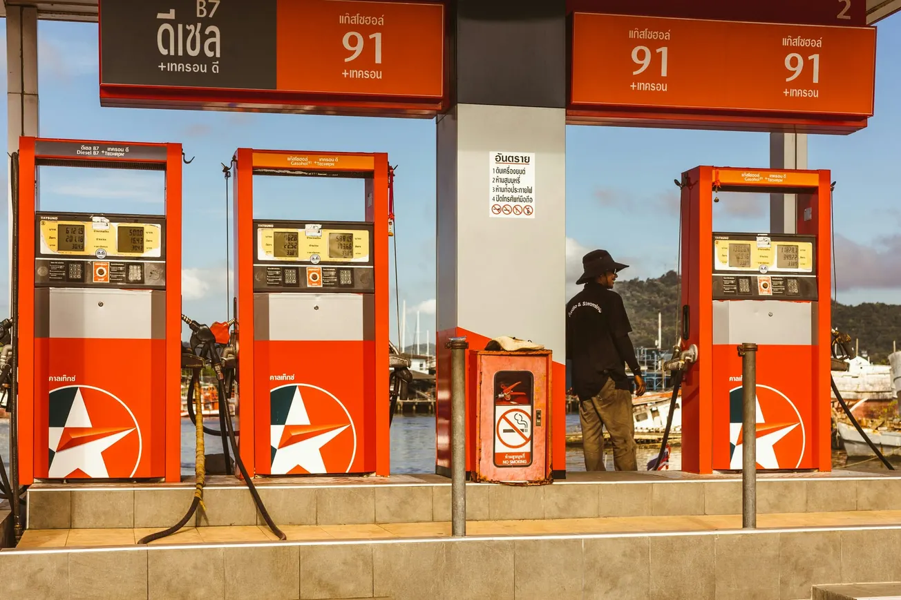Three red gas pumps stand in a row at a station near a waterfront. A person in a hat and dark clothes is using the rightmost pump. Bright, sunny day.
