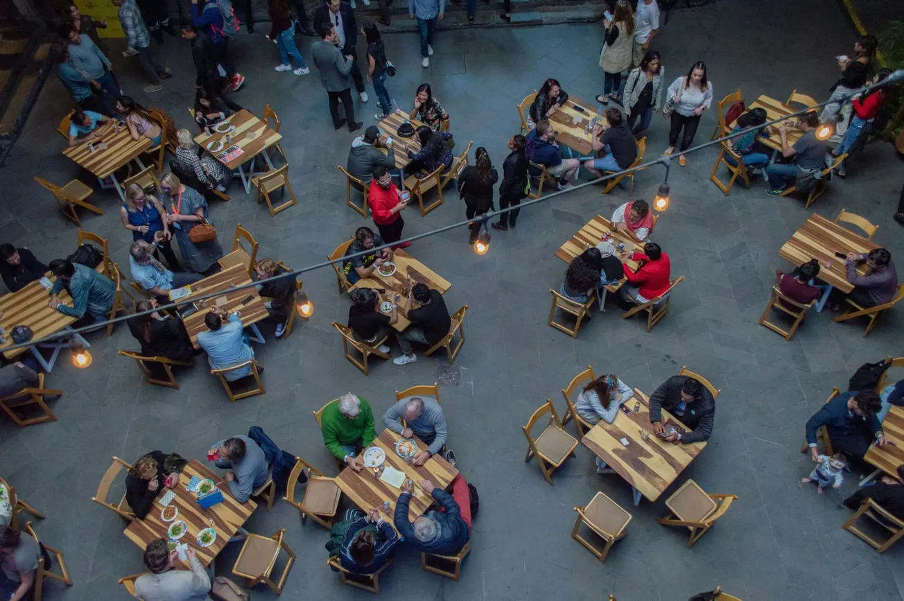 Aerial view of a bustling café with wooden tables and chairs, people socializing and dining. Warm lights create a cozy atmosphere.