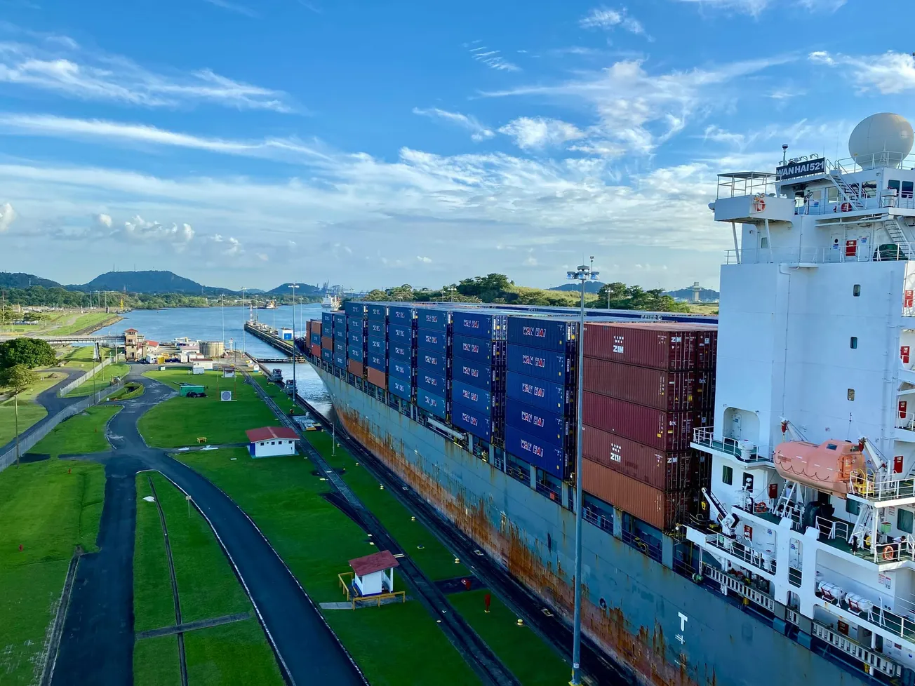 A large container ship travels through a canal under a bright blue sky, flanked by green grassy banks with scattered small buildings.