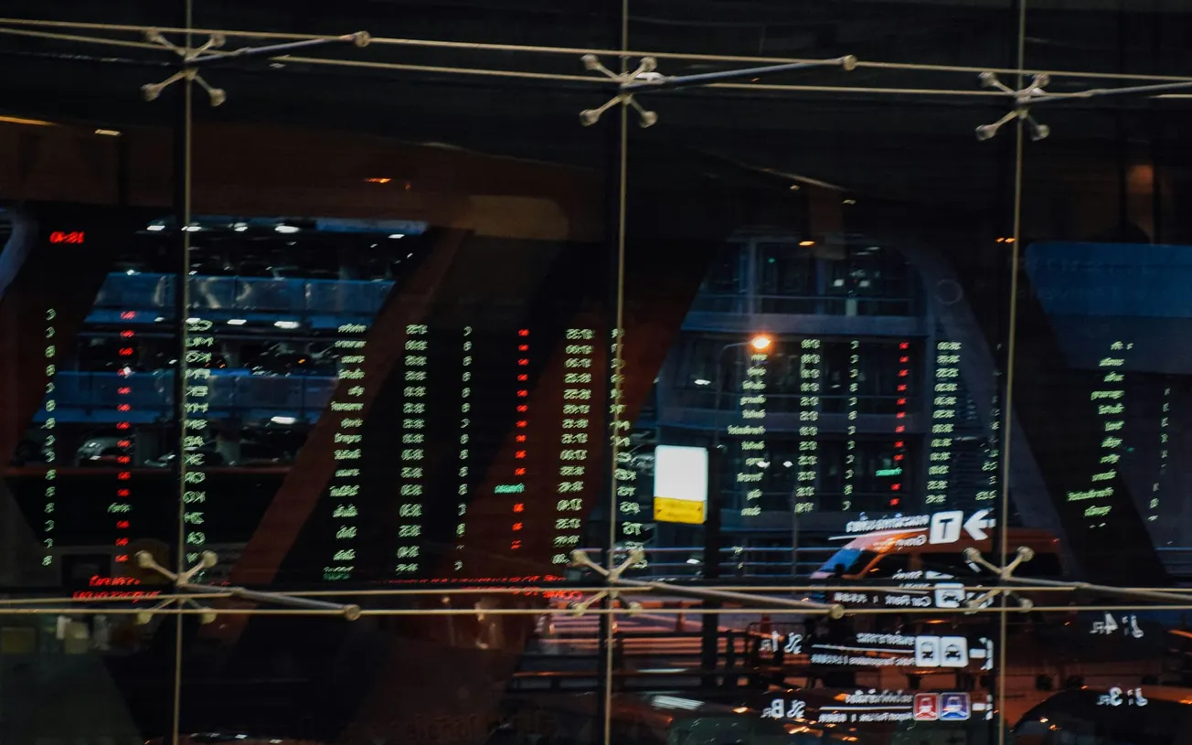Reflection of an airport departure board on glass panels, displaying flight details in glowing red and green text. The scene is bustling and dynamic.