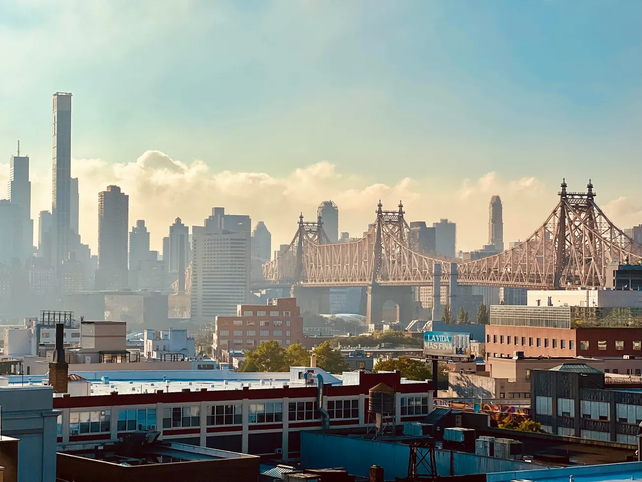 View of a city skyline with tall skyscrapers against a foggy sky and a prominent suspension bridge. Foreground includes industrial buildings, conveying urban vibrancy.
