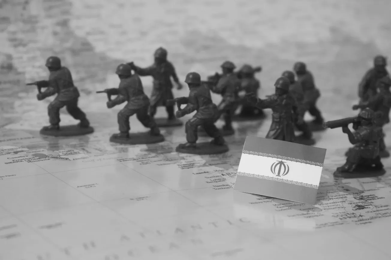 Toy soldiers carrying guns are displayed on a map next to a small Iranian flag, suggesting a military strategy or conflict theme. The image is in black and white.