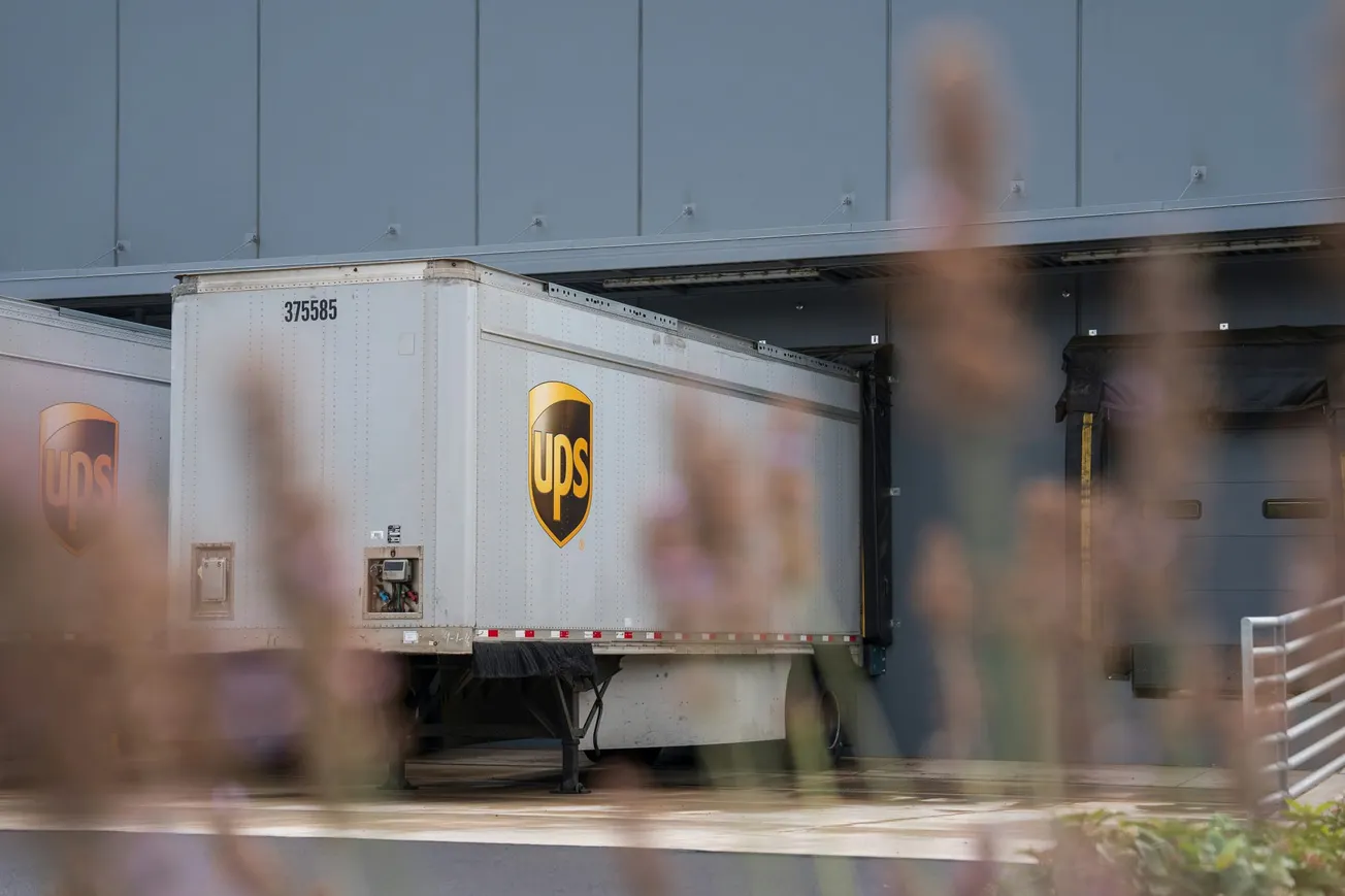 A UPS trailer backed into a large loading dock at an industrial building, partially obscured by out-of-focus plants in the foreground, conveys a busy, industrial feel.