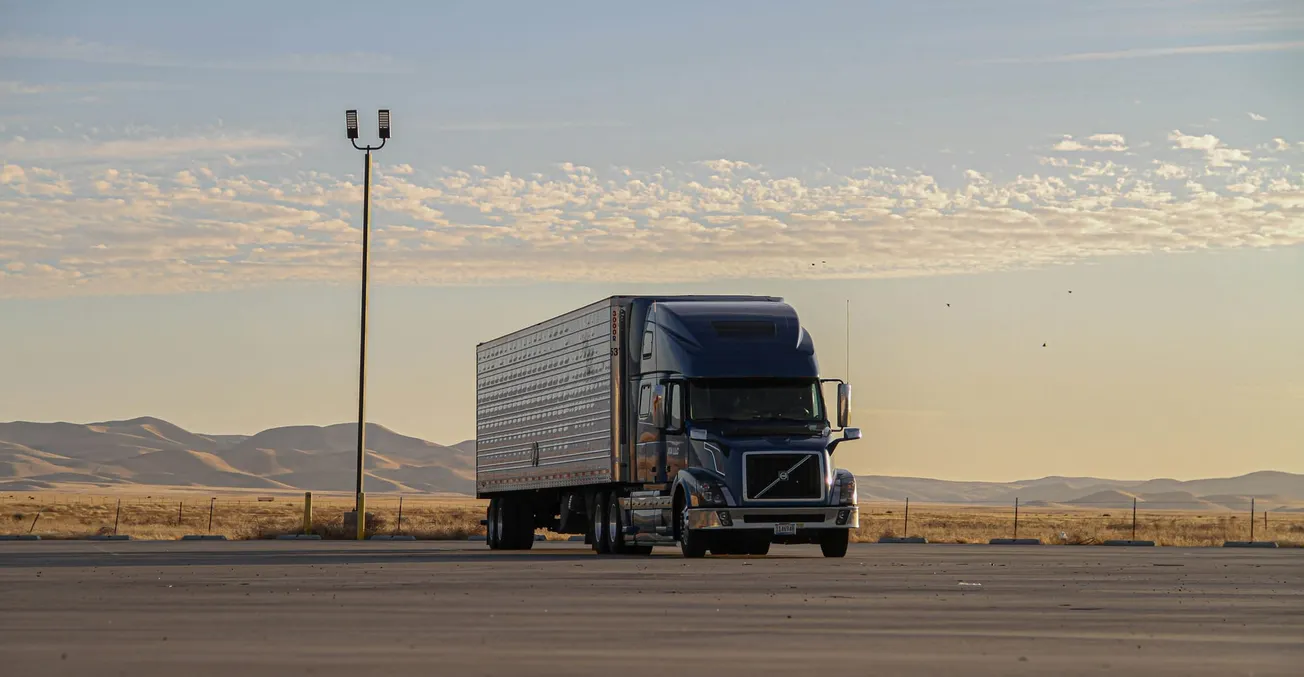 A blue semi-truck parked in a vast, empty lot under a pale sky with scattered clouds. Distant mountains create a serene and expansive backdrop.