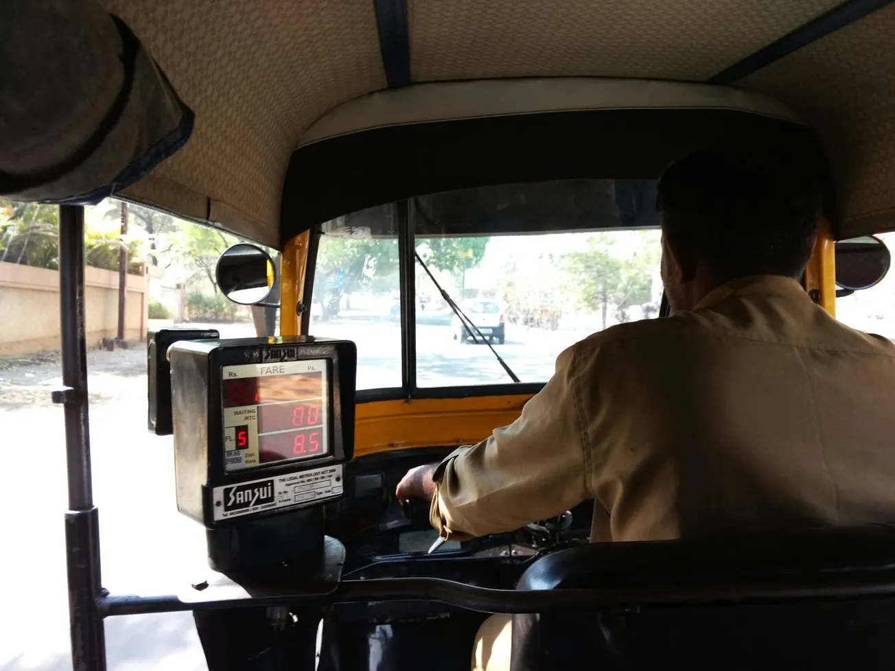 Interior view of a moving auto-rickshaw, showing the driver in a beige shirt and a fare meter reading 0.0. Sunlight filters through, casting shadows.