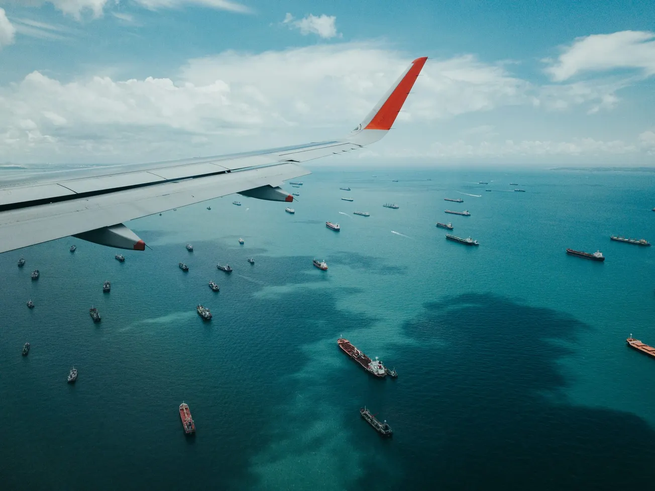 Aerial view of a wing from an airplane flying above a sea dotted with numerous ships. The sky is clear with a few clouds, conveying a serene atmosphere.