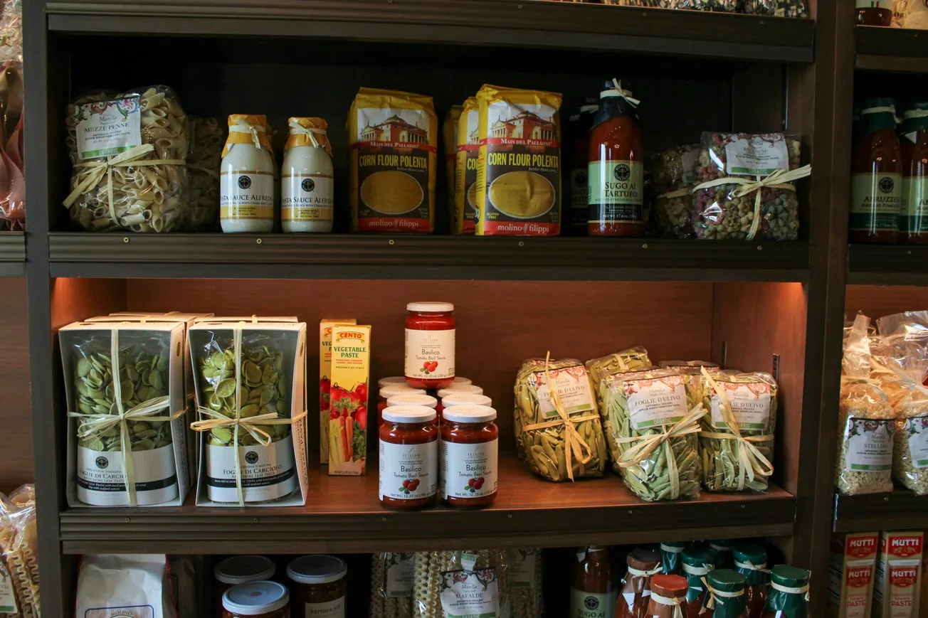 Wooden shelves displaying a variety of packaged Italian foods, including pasta, corn flour, and tomato sauce. The setup evokes a cozy Italian deli.