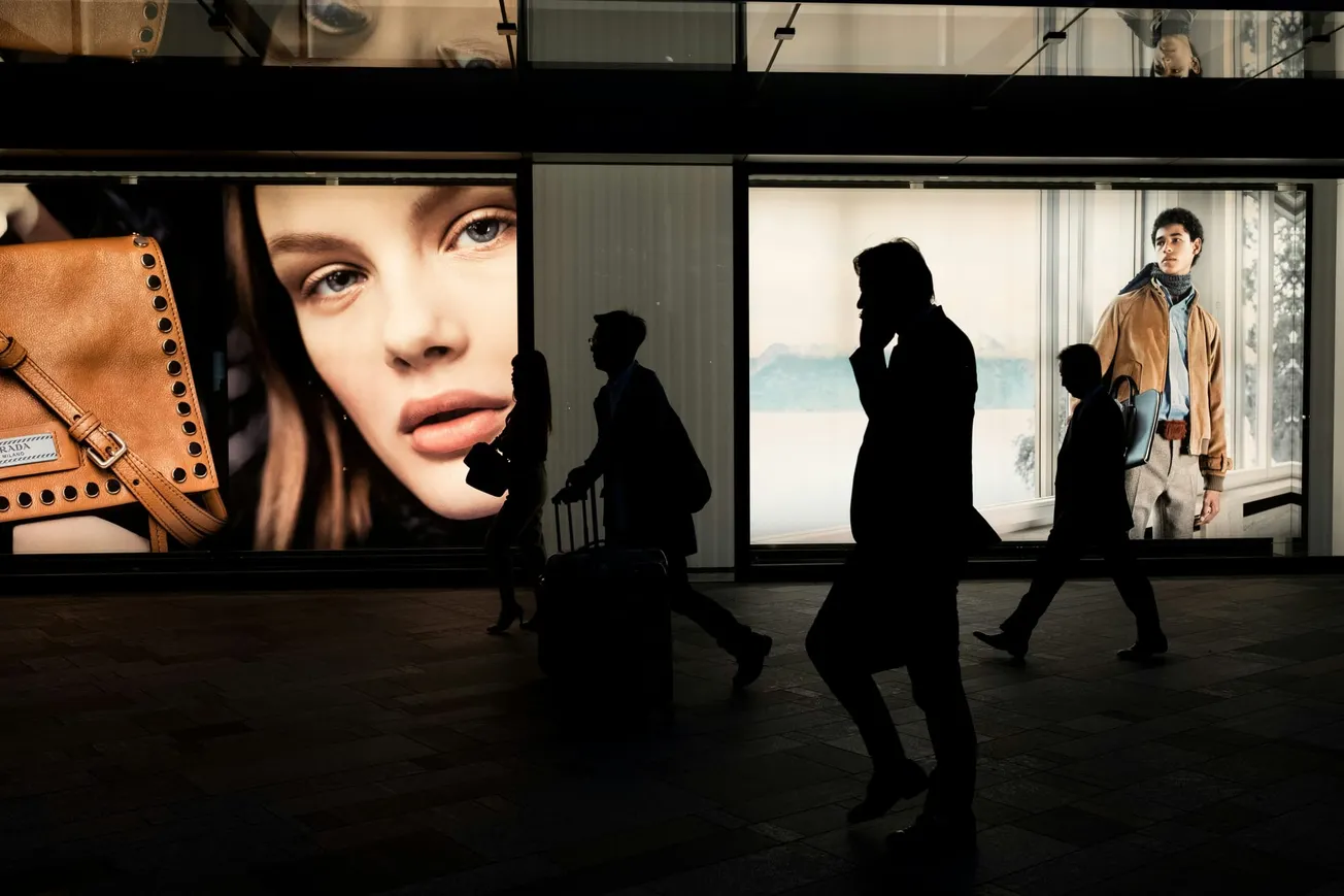 Silhouettes of people walk past large fashion ads in a shopping area. The ads show a close-up of a woman's face and a man in a jacket, conveying a stylish urban vibe.