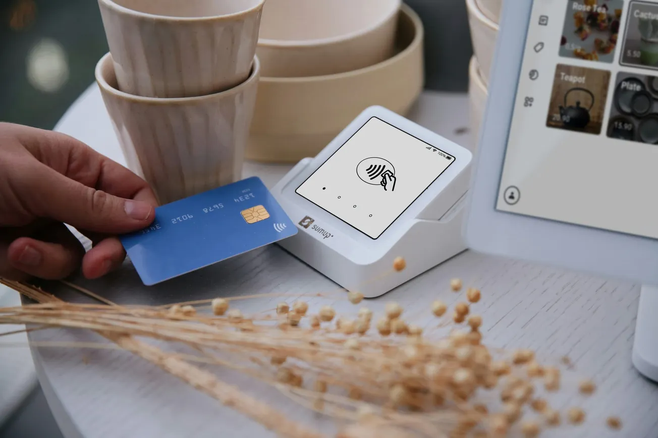 A person taps a blue contactless credit card on a white payment terminal. In the background, there are stacked cups and a tablet screen displaying product images.