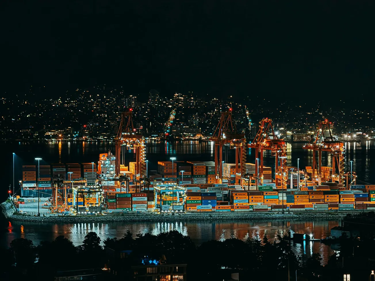 Nighttime view of a bustling port with large cranes and stacked shipping containers illuminated by bright lights, against a city skyline backdrop.