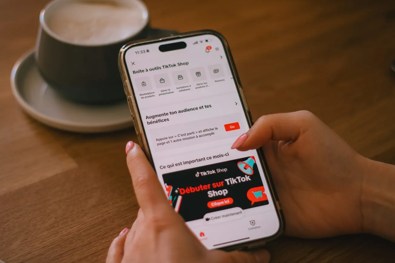 Hands holding a smartphone displaying TikTok Shop interface on a wooden table, with a cup of coffee nearby. The tone is casual and focused.