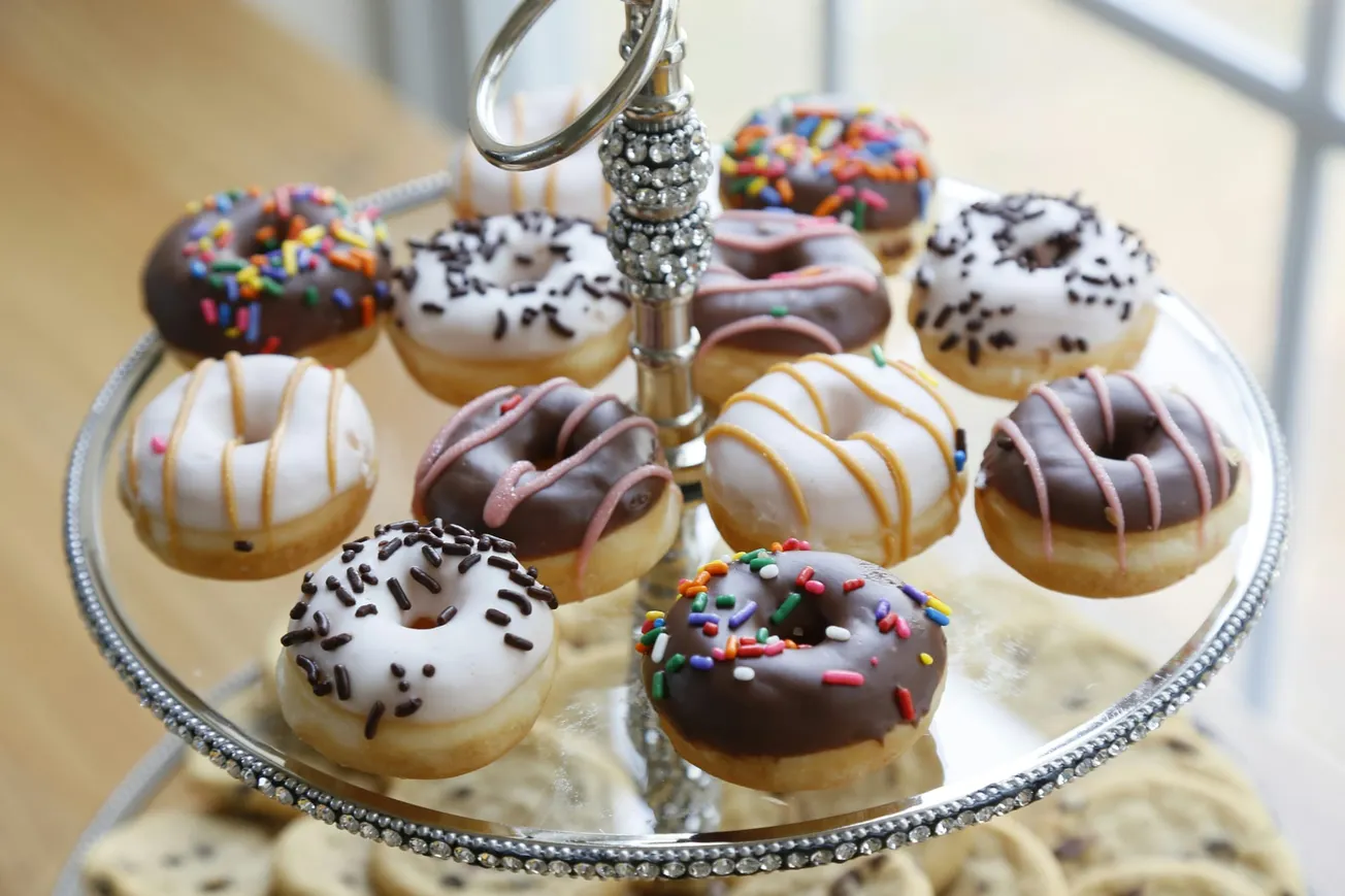 A silver tiered tray holds an assortment of mini donuts with colorful toppings. Frostings include white and chocolate with sprinkles and drizzles. Bright and inviting.
