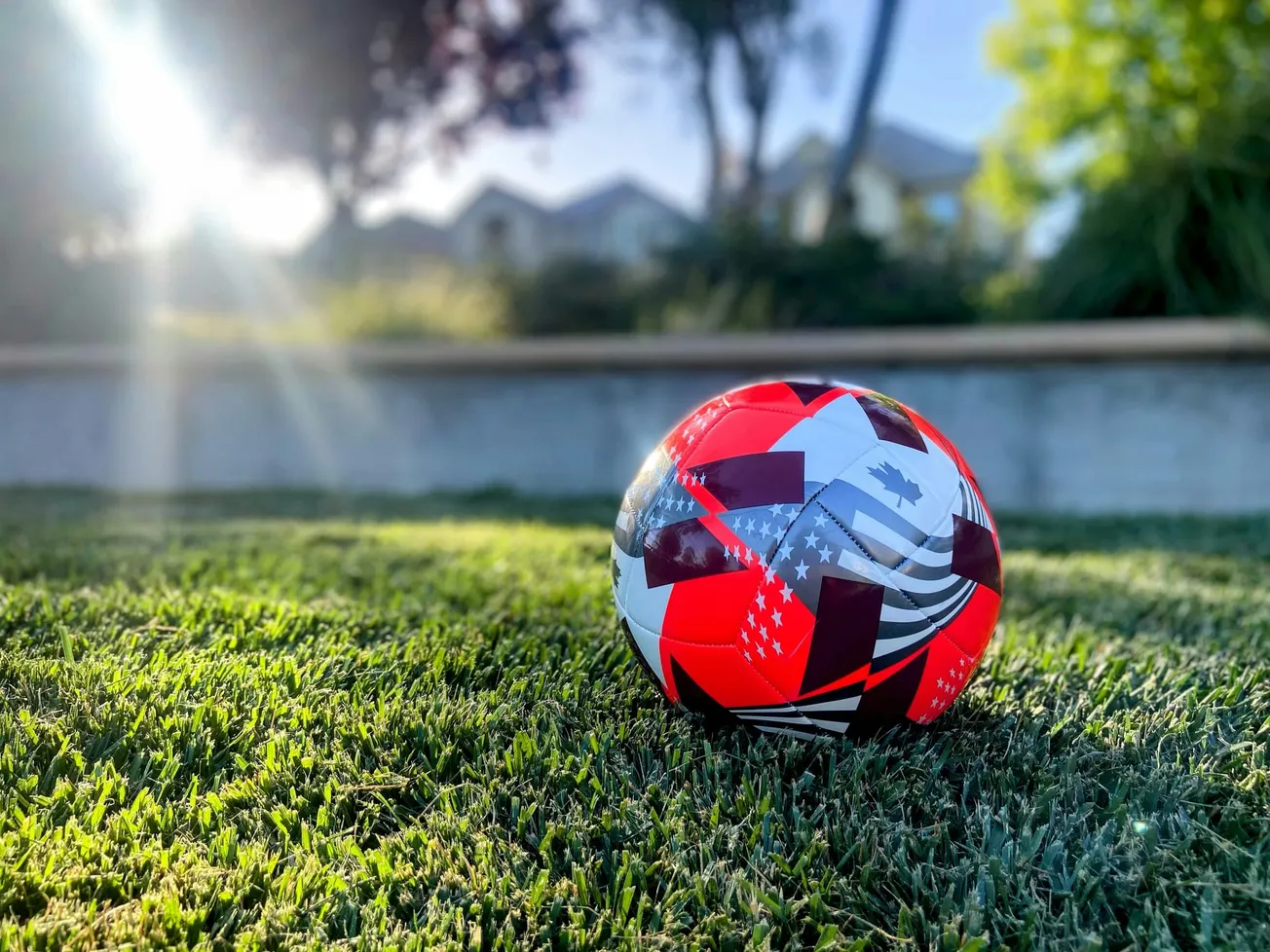 A vibrant red soccer ball marked with geometric patterns and star designs rests on sunlit grass. Blurred background with trees and houses.