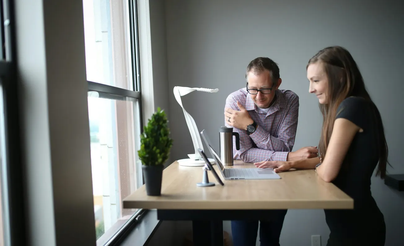 Two people collaborating at a standing desk, focused on a laptop. A large window and potted plant create a bright, modern office atmosphere.