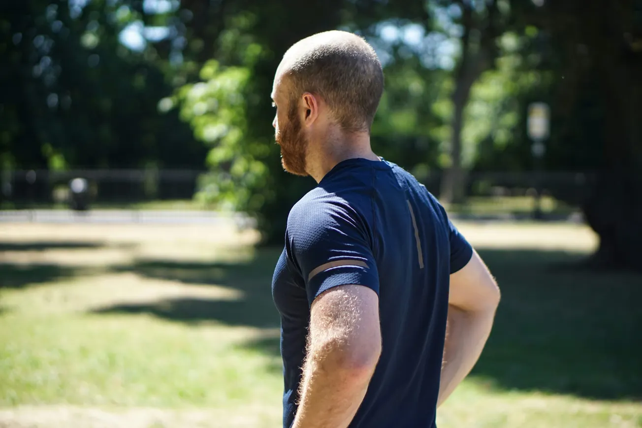 A man in a blue athletic shirt stands with hands on hips in a sunny park, surrounded by greenery. His posture suggests a moment of rest or contemplation.