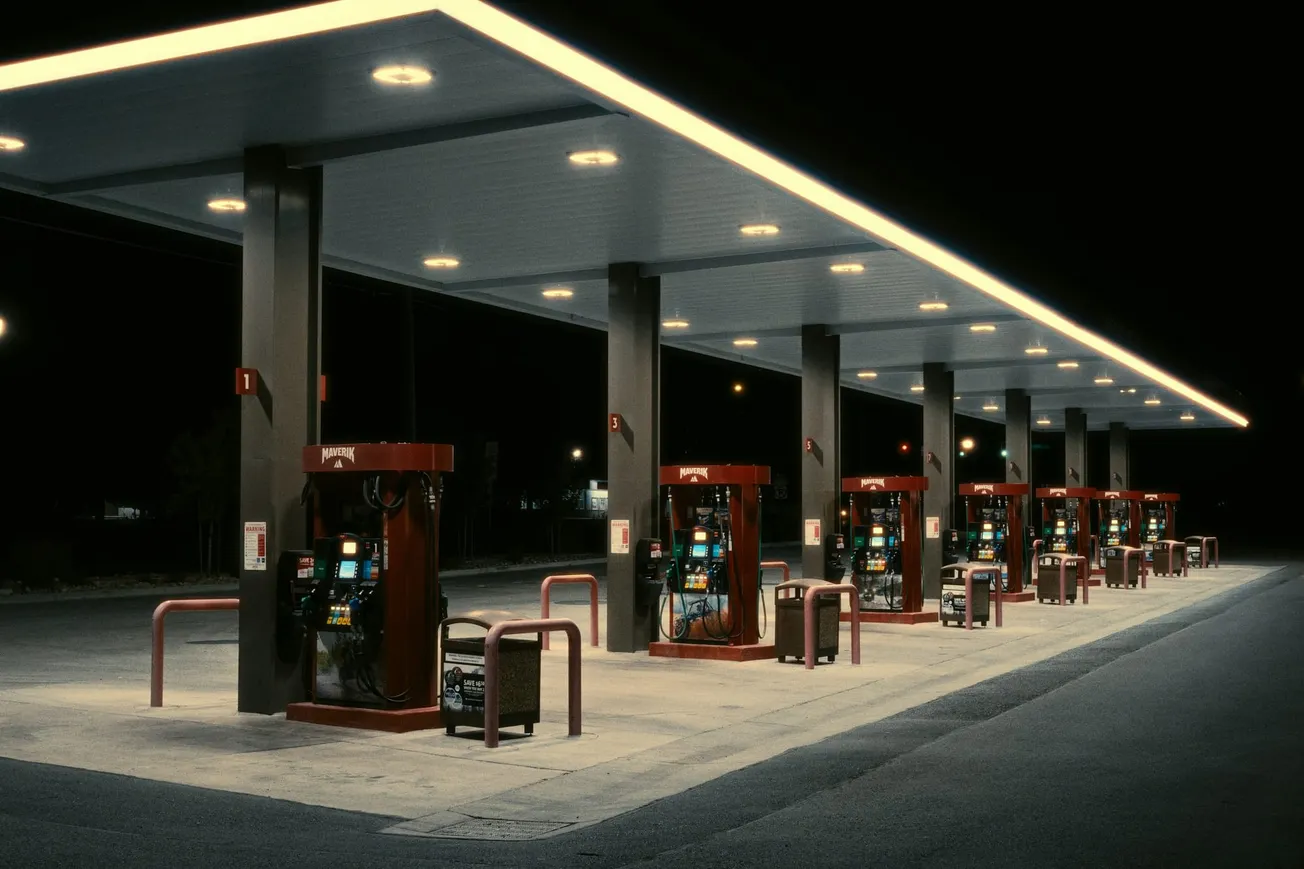 A brightly lit gas station at night, featuring a row of red fuel pumps under a wide canopy. The area is empty, creating a quiet and isolated atmosphere.