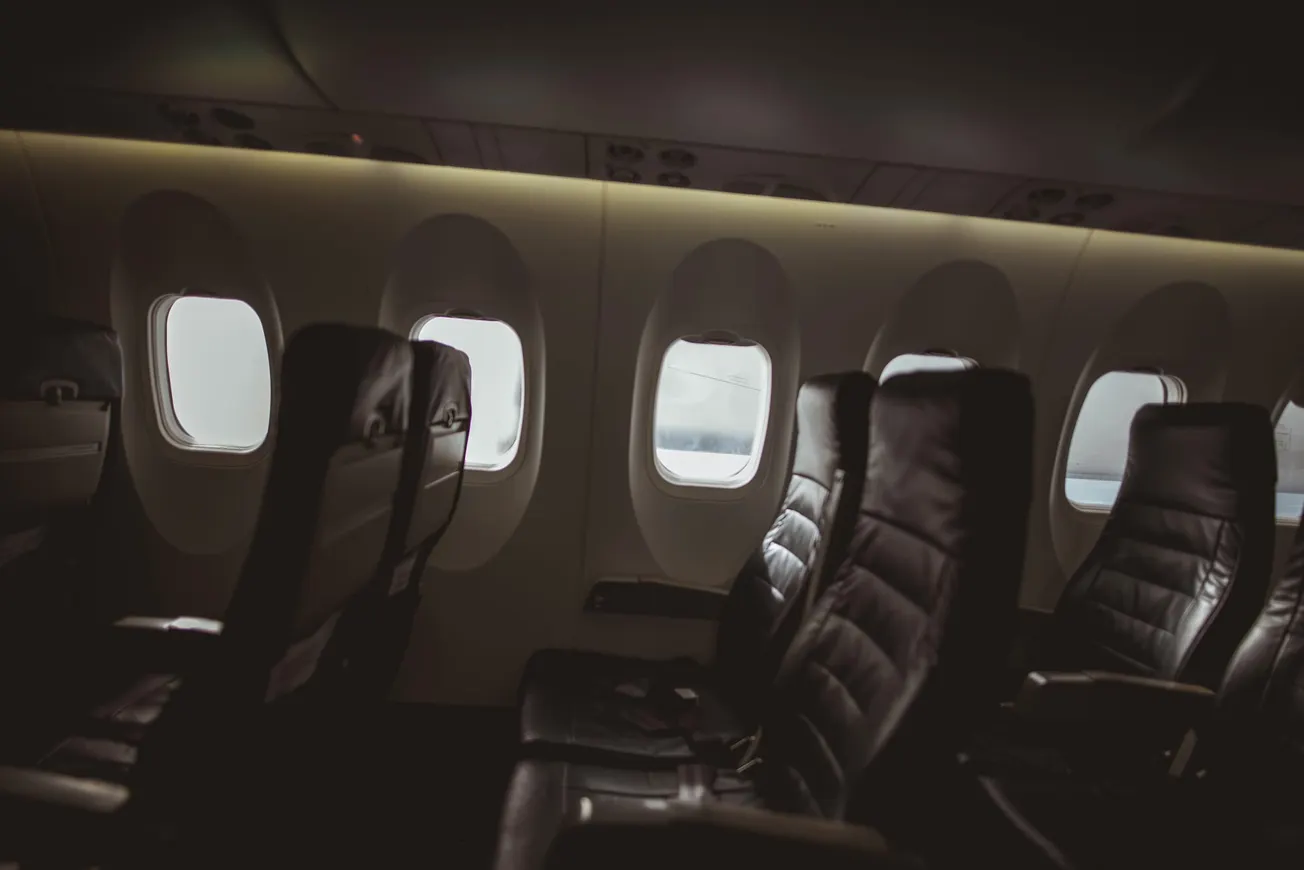Empty airplane seats in dim lighting, aligned next to oval windows showing a cloudy sky, convey a calm and quiet atmosphere.