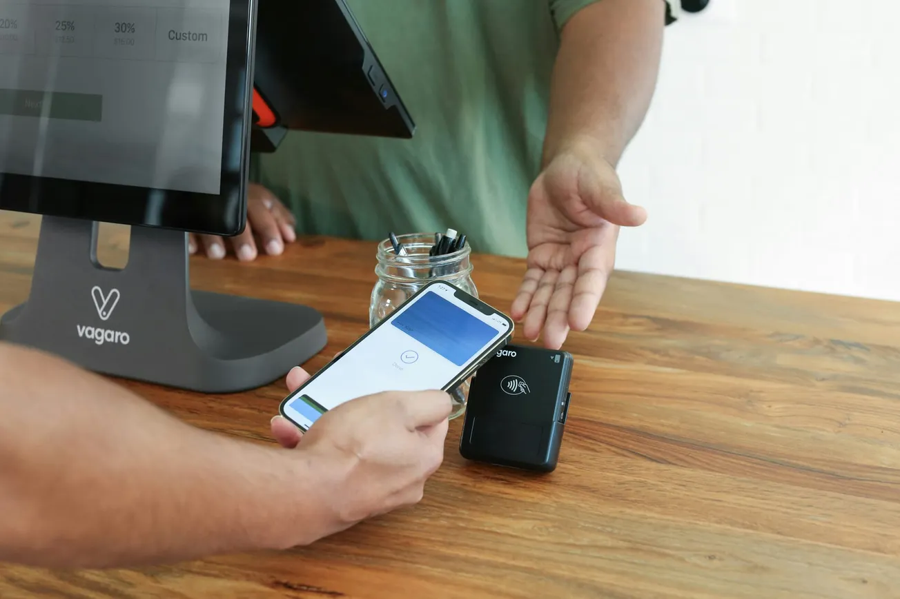 Person making a contactless payment with a smartphone at a point-of-sale terminal on a wooden counter. Another person gestures towards the card reader.