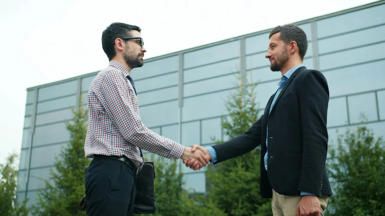 Two men in professional attire shake hands in front of a modern glass office building and evergreen trees.