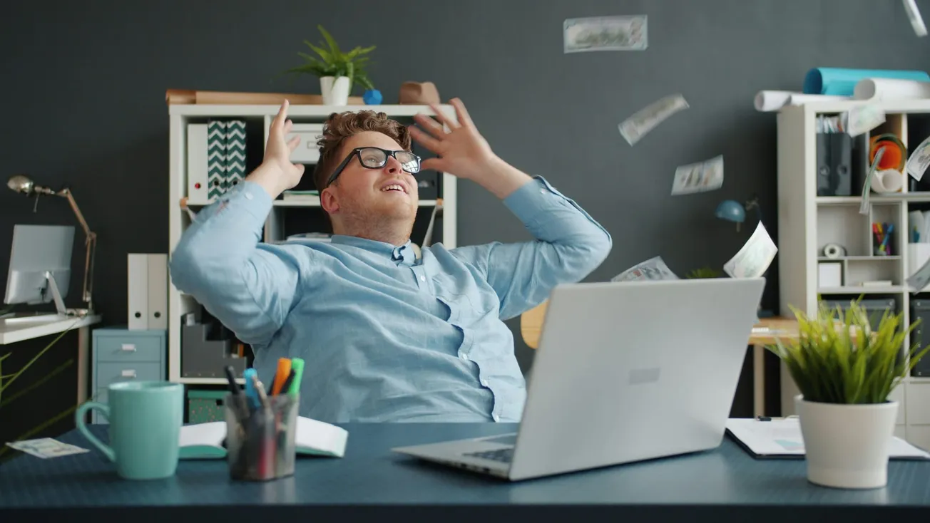 Man in a blue shirt joyfully leans back in an office chair, throwing money into the air. A laptop and office supplies are on the desk, conveying a celebratory mood.