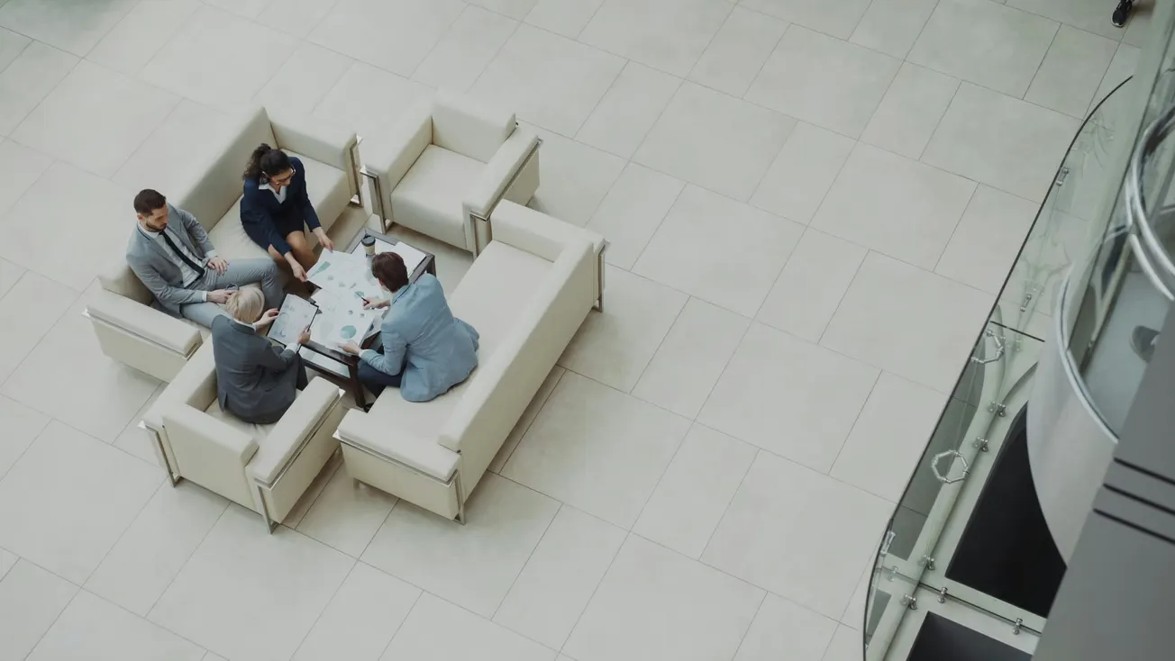 Overhead view of four business professionals engaged in a meeting at a table surrounded by cream-colored sofas, set in a spacious, modern lobby.