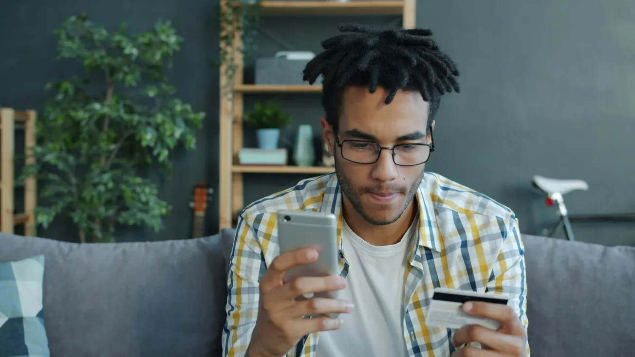 A man with glasses, wearing a plaid shirt, looks focused while holding a smartphone and a credit card. He's seated on a couch, with shelves and plants in the background.
