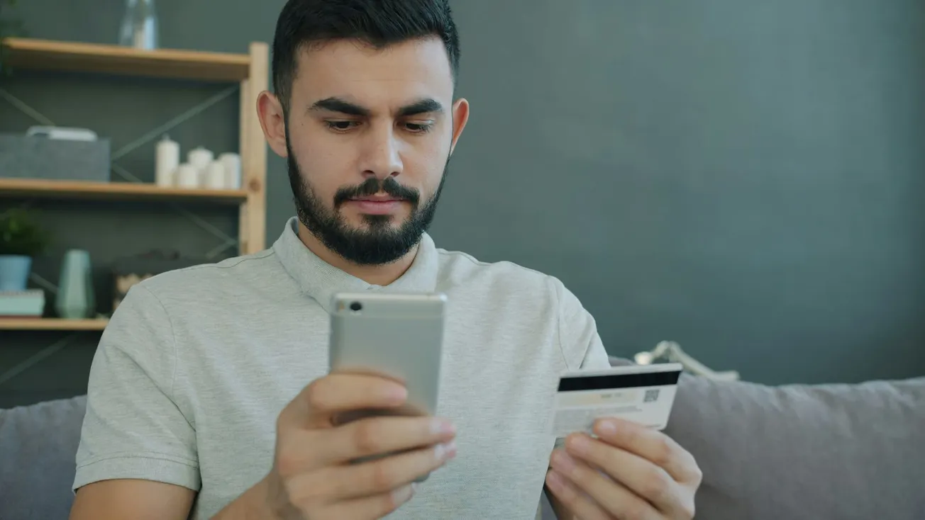 A man with a beard sits on a couch, focused on his smartphone, holding a credit card. The room is modern, with shelves and decorations in the background.
