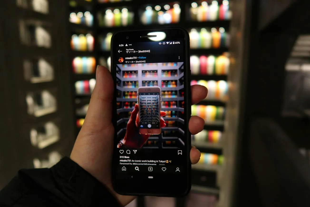 A hand holds a smartphone displaying an Instagram post, capturing a similar image of colorful bottles on shelves in the background, creating a visual loop effect.