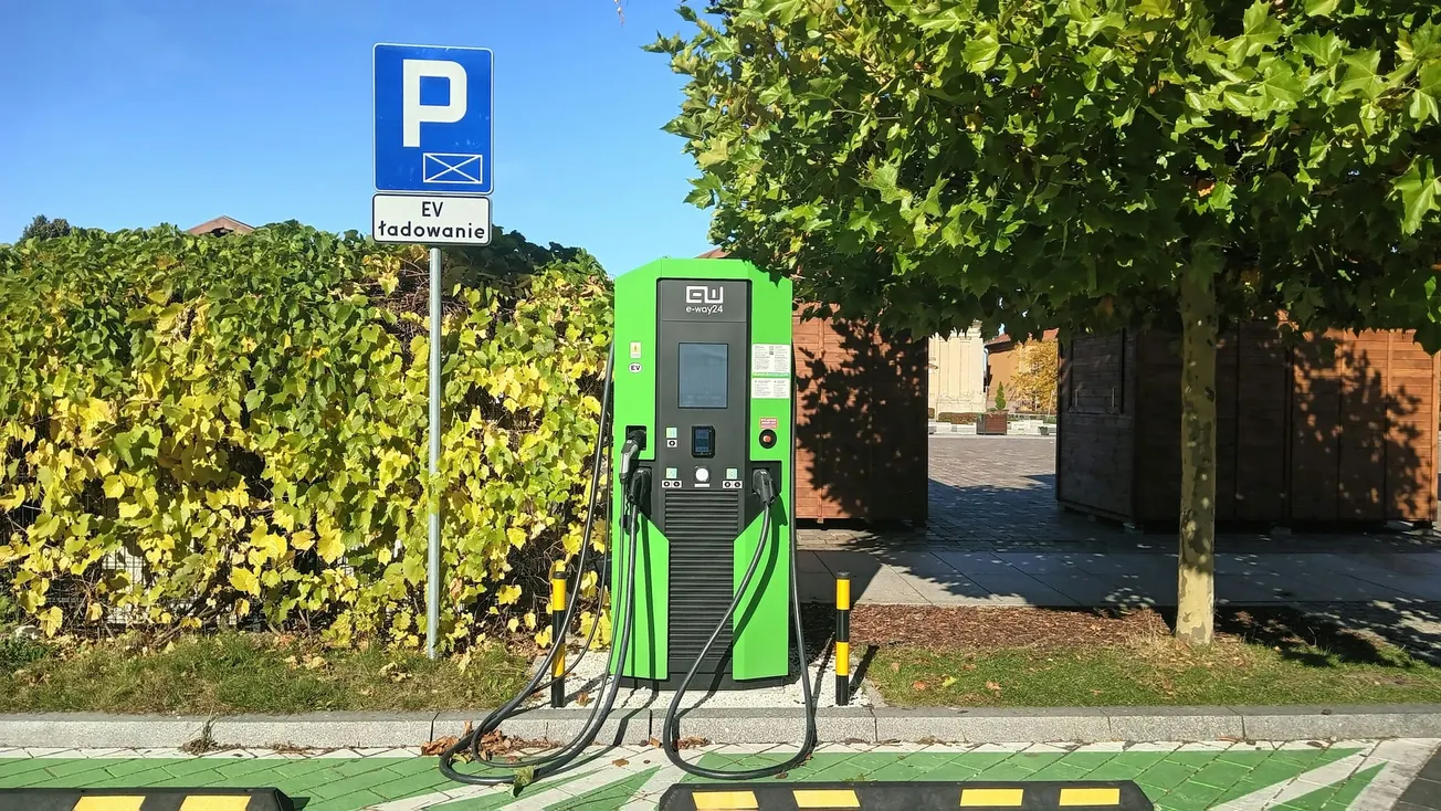 Electric vehicle charging station in a parking area with a "P" sign above labeled "EV ładowanie." Green foliage and a tree are in the background.