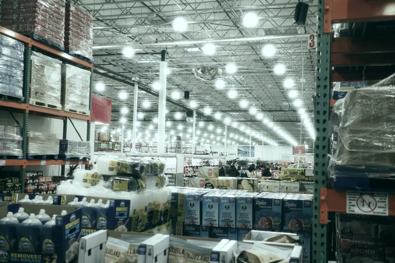Wide-angle view of a large warehouse store filled with shelves of bulk packages under bright ceiling lights. The atmosphere is bustling and organized.