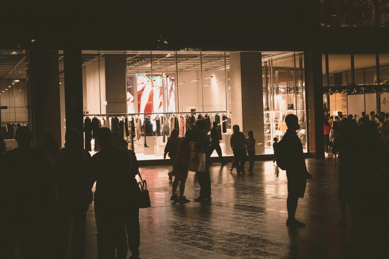 Shoppers stroll past a brightly lit clothing store with large windows displaying racks of clothes, casting silhouettes and reflections on the wet pavement.