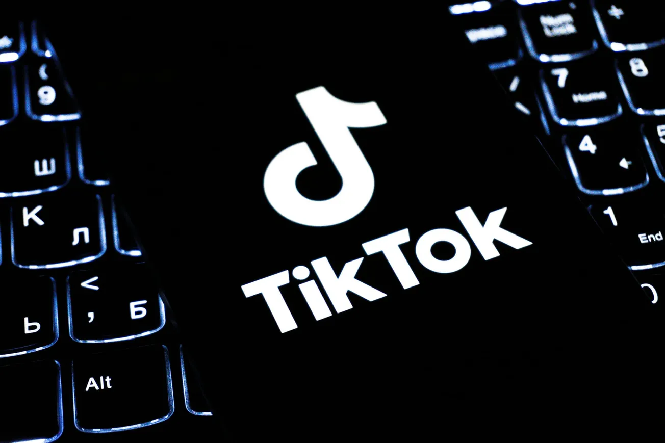 A smartphone displaying the TikTok logo rests on a dimly lit keyboard. The glow from the screen and keys creates a tech-focused, modern ambiance.