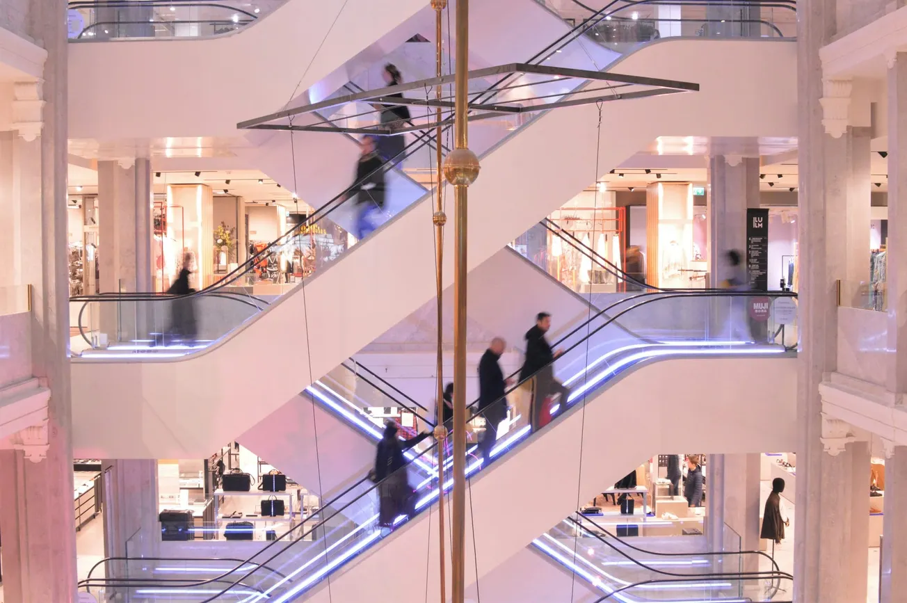 Shoppers on multiple levels of modern escalators in a brightly lit department store. The scene conveys a busy, bustling atmosphere.