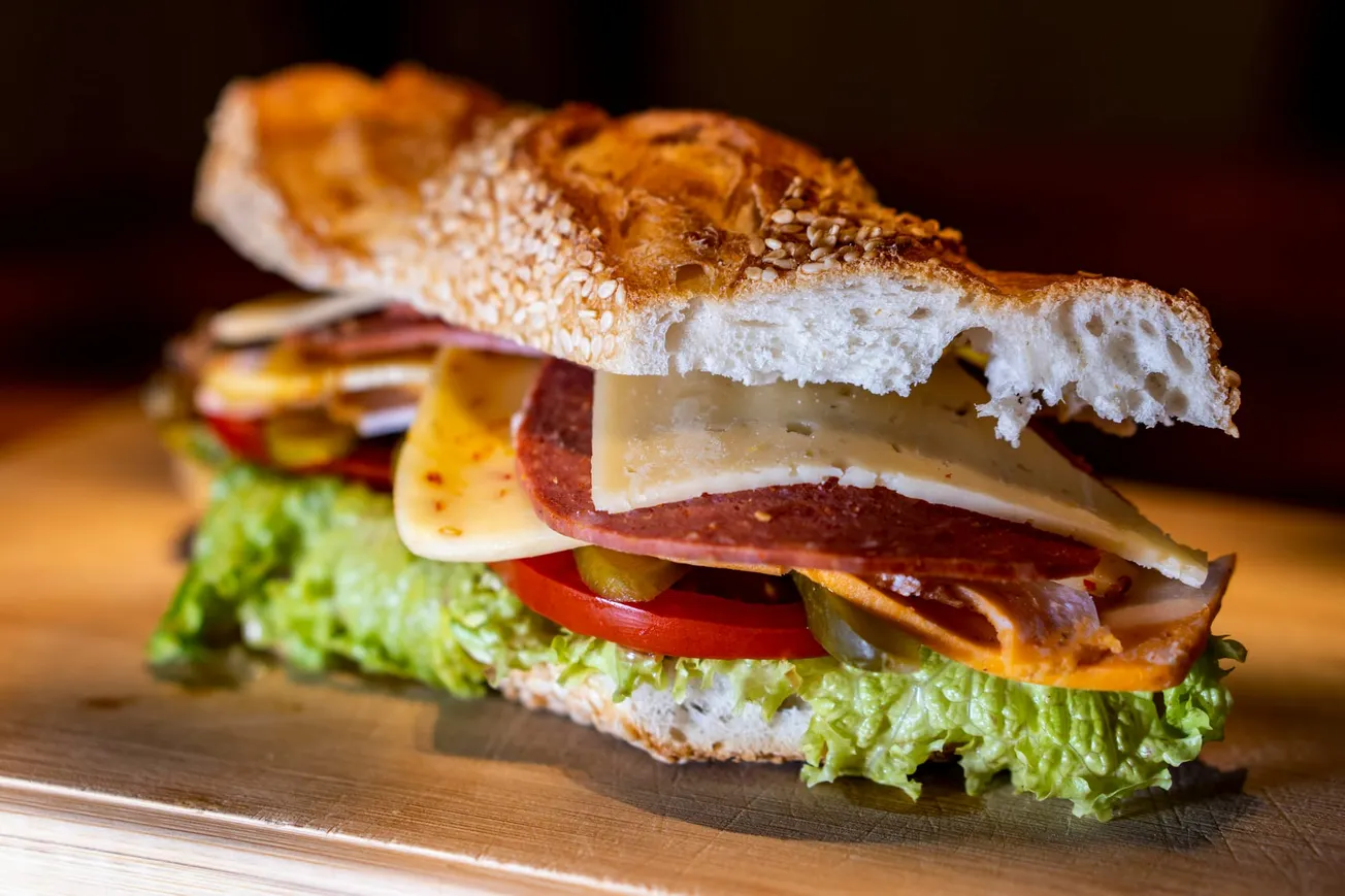 A close-up of a fresh sandwich with sesame-crusted bread, layered with lettuce, tomato, cheese, salami, and pickles, set on a wooden surface.