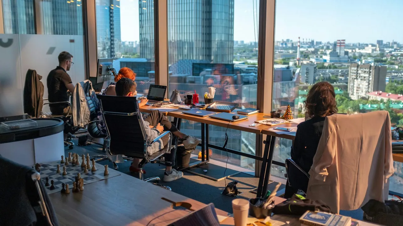 Office with four people working at desks, laptops in use. Large windows showcase a cityscape outside. Chess set on a table suggests strategy or leisure.