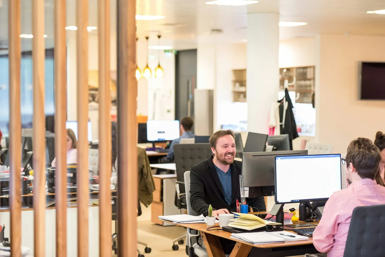 Open-plan office with people working at desks. A person in a black blazer smiles while talking to a colleague in a pink shirt.