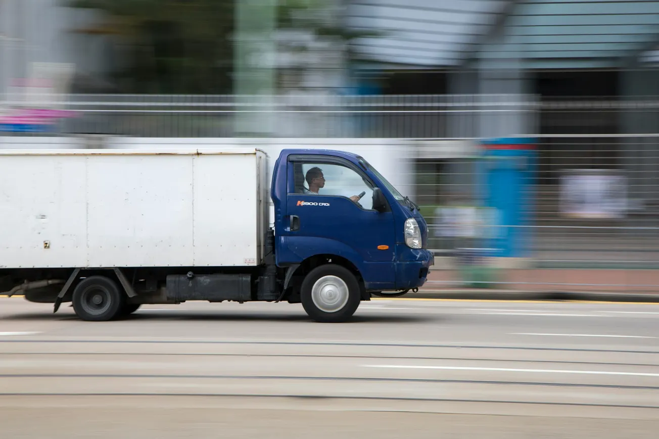 A blue and white delivery truck speeds down a city street, creating a blurred motion effect. The driver is visible, focused on the road ahead.